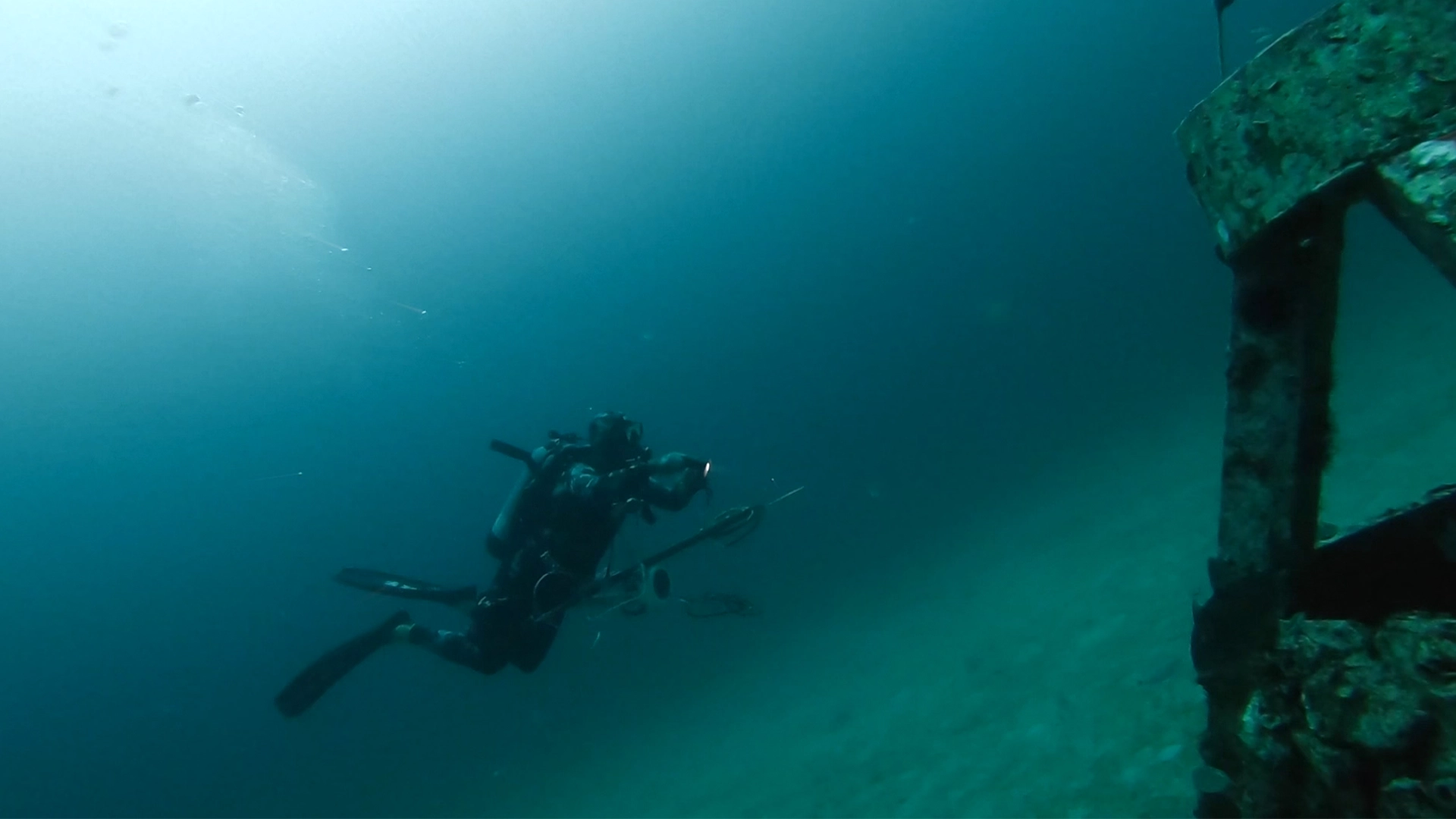 Scuba diver with a speargun swimming near an underwater structure in deep blue water.